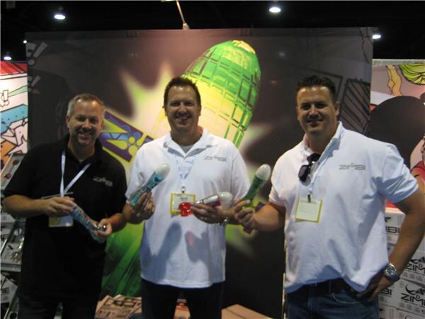 Three men with badges hold colorful Zimbi products in front of a booth backdrop with a green rocket graphic.