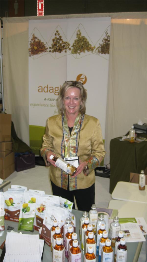 A smiling woman with sunglasses on her head and a yellow jacket holds a bottle at the Adagio booth, with tea bags and bottled teas on the table and a banner of loose tea behind her.