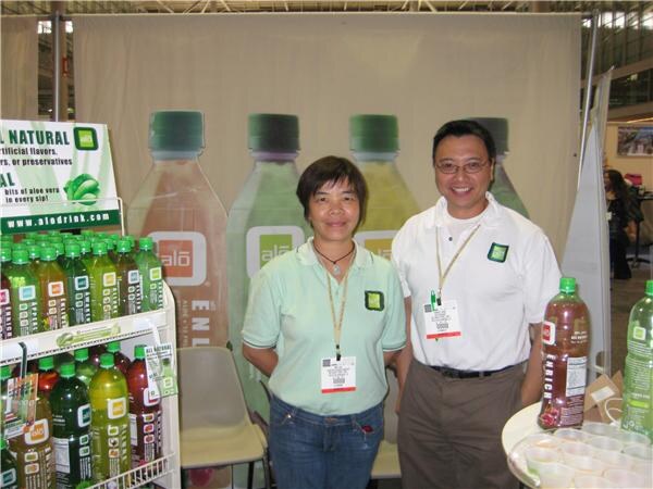 A woman in a light green shirt and a man in a white polo, both wearing badges, stand together at an Alo booth with bottles of aloe drink on a shelf and large bottle graphics behind them.