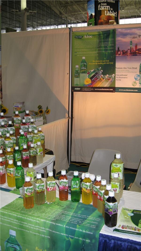 Booth table crowded with bottles of Aloe Farm aloe vera drink in various colors, with a green Aloe Farm banner behind them and a chair against a white curtain.