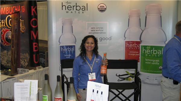 A smiling woman with curly dark hair in a blue shirt holds an orange-labeled bottle in front of Ayala's Herbal Water banners, with more bottles on the table in front of her.
