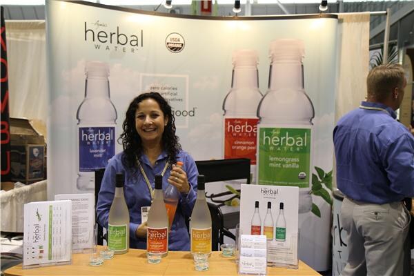 A smiling woman in a blue shirt holds a bottle at a table of Ayala's Herbal Water bottles and flyers, with a man in a blue shirt beside her and large herbal water banners behind.
