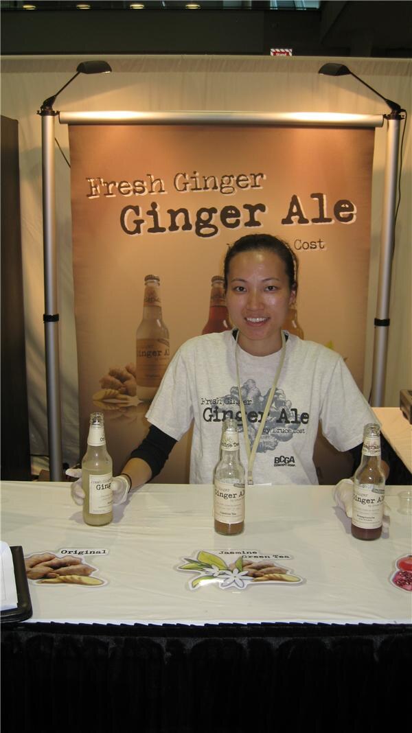 A smiling woman in a Fresh Ginger Ginger Ale T-shirt stands behind a table with three bottles of ginger ale, in front of a Bruce Cost Fresh Ginger Ginger Ale banner.