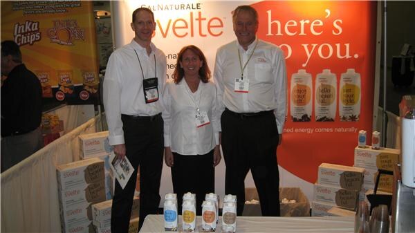 Two men and a woman in white shirts with badges stand at the Cal Naturale Svelte booth, with cartons and bottles on the table and an Inka Chips sign at left.
