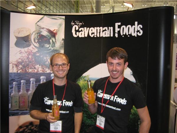 Two smiling men in black Caveman Foods t-shirts and red lanyards stand in front of a Caveman Foods display, each holding a bottle of orange-colored drink.