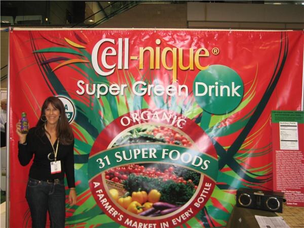 A woman with long dark hair holds a small bottle beside a large red Cell-nique Super Green Drink banner reading Organic, 31 Super Foods, A Farmer's Market in Every Bottle.