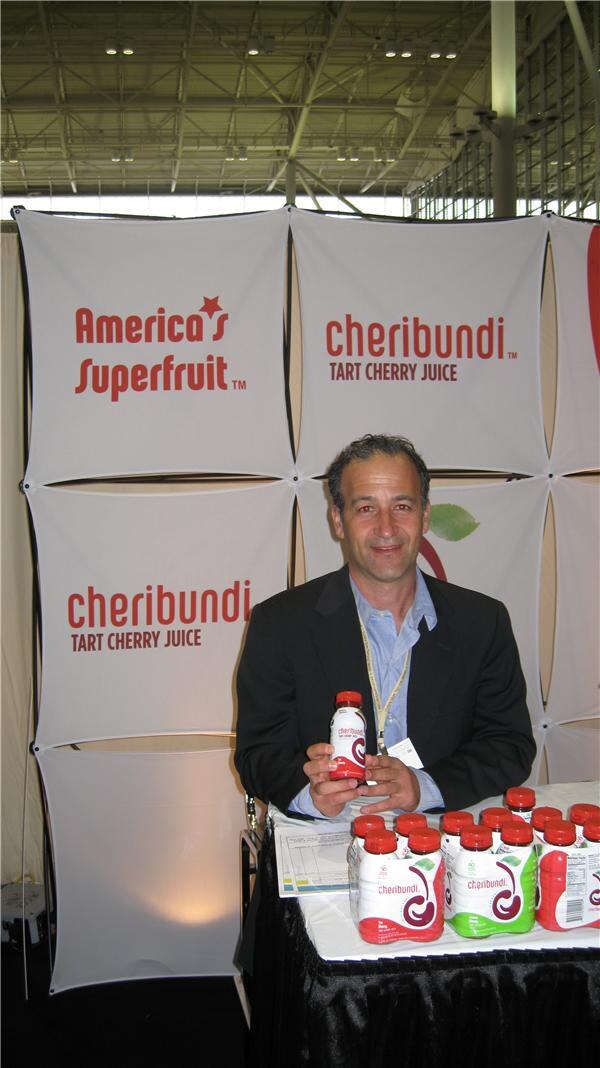A man in a dark jacket and light blue shirt sits at a table holding a Cheribundi tart cherry juice bottle, with more bottles on the table and Cheribundi banners behind him.