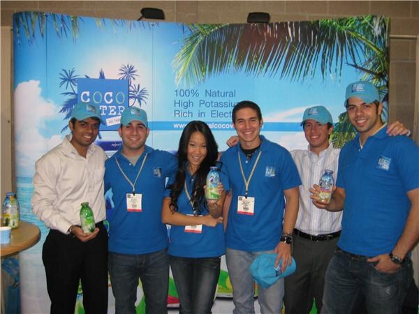 Six people, most in blue Coco Water shirts and caps, pose together in front of a tropical Coco Water backdrop, several holding bottles of coconut water.