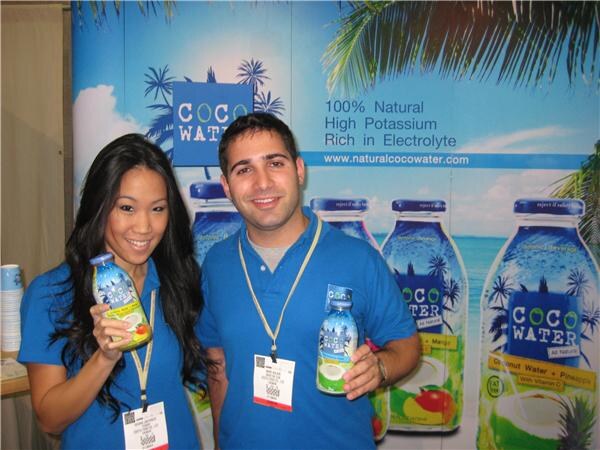 A woman and a man in blue shirts and lanyards smile and hold small Coco Water bottles in front of a backdrop showing large Coco Water bottles and palm trees.