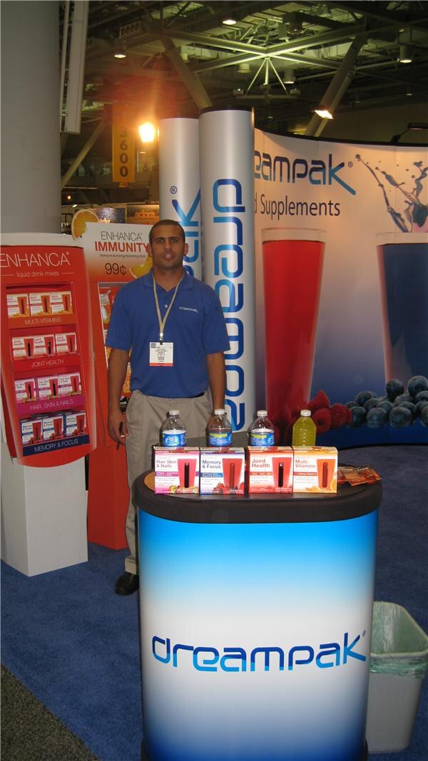 A man in a blue polo stands behind a Dreampak counter holding boxed Enhança liquid drink mixes and water bottles, with an Enhança display rack and Dreampak banners beside him.
