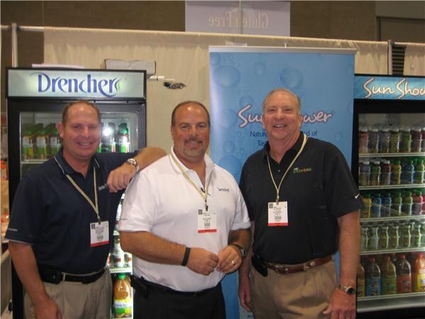 Three men in polo shirts and lanyards stand between refrigerated coolers labeled Drencher and SunShower, which are stocked with bottled drinks.