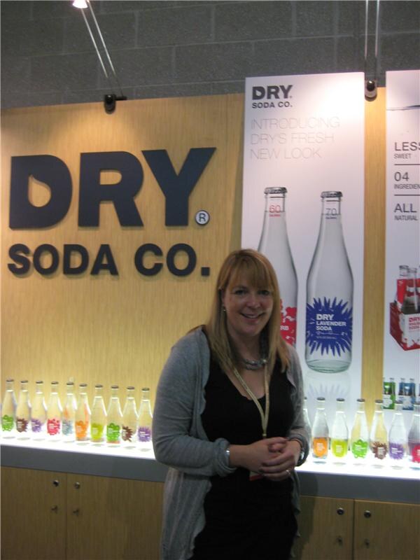 A smiling woman with a lanyard stands in front of a Dry Soda Co. sign and a lit shelf of colorful glass soda bottles, beside a poster showing Dry Lavender Soda.