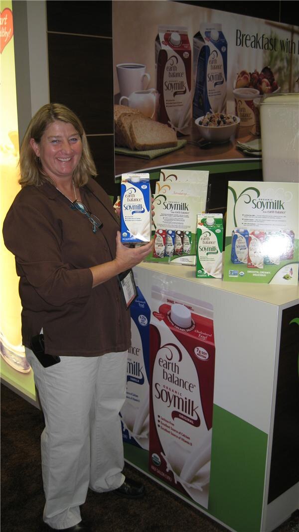 A smiling woman in a brown top holds a carton of Earth Balance Organic Soymilk beside a display of soymilk cartons and signs at a trade show booth.