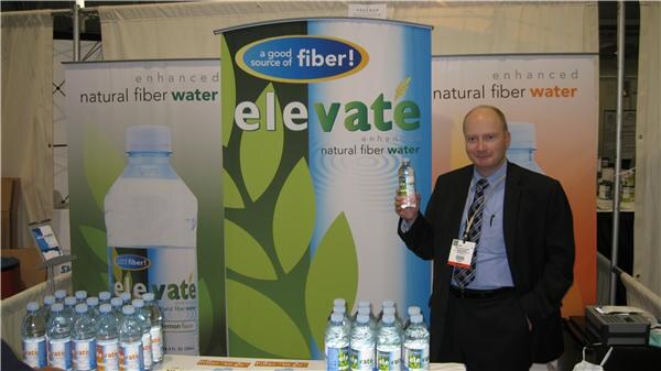 A man in a dark suit holds a bottle of Elevate enhanced natural fiber water beside Elevate banners, with more bottles lined up on the table in front.