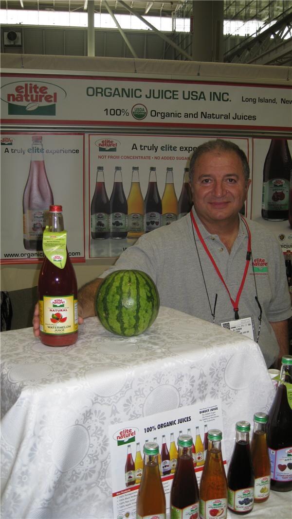 A smiling man in a gray polo stands behind a table with a watermelon and a bottle of Elite Naturel watermelon juice, under an Organic Juice USA Inc. sign.