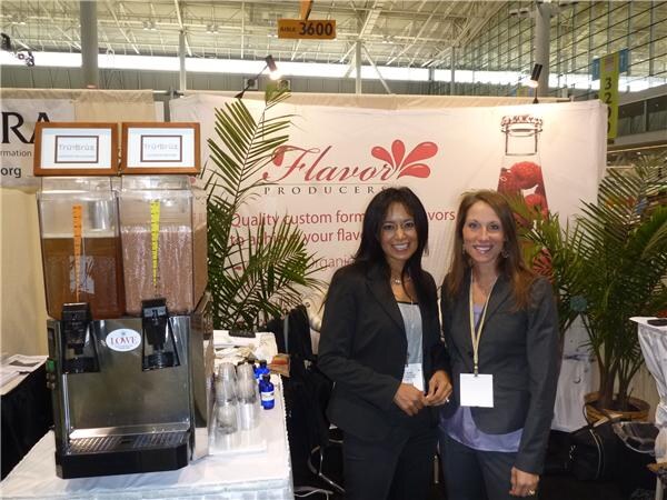 Two smiling women in dark blazers stand at a Flavor Producers booth next to a beverage dispenser with two tanks, in front of a banner with the company name.