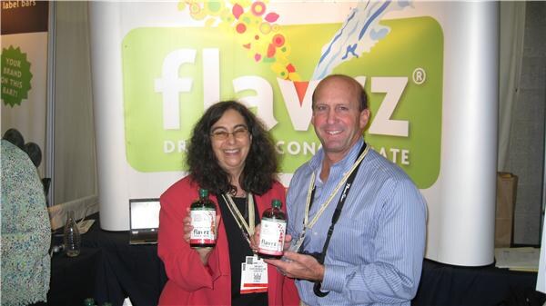 A woman in a red jacket and a man in a blue striped shirt each hold bottles of Flavrz in front of a green and white Flavrz sign.