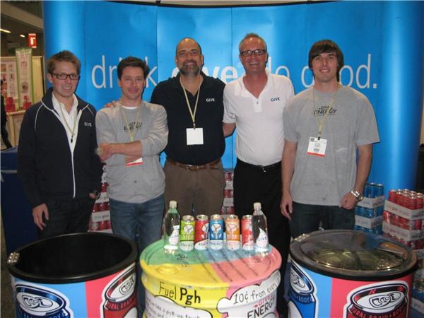 Five men stand in a row behind a table of GIVE water and energy drink cans and bottles, in front of a blue banner with a drink-themed slogan.