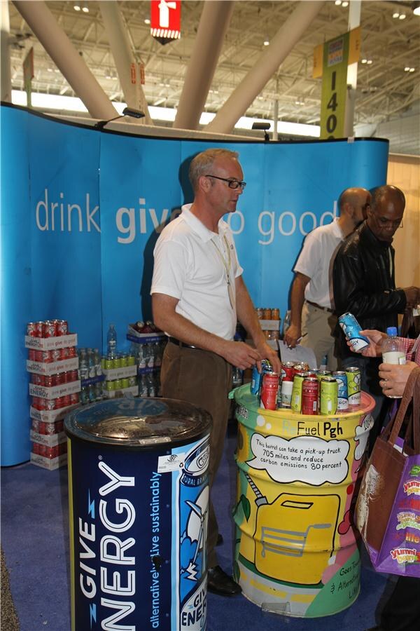 A man in a white polo stands at a blue Give booth with a Give Energy barrel cooler and a painted barrel holding cans, as other attendees stand nearby.