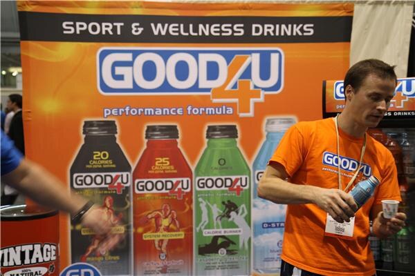 A man in an orange Good4U shirt holds a bottle and a small cup in front of a Good4U sport and wellness drinks banner showing four bottle flavors.