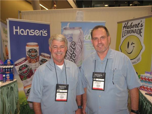 Two men in light blue shirts and Hansen's badges stand in front of Hansen's can banners, with a Hubert's Lemonade sign to the right.