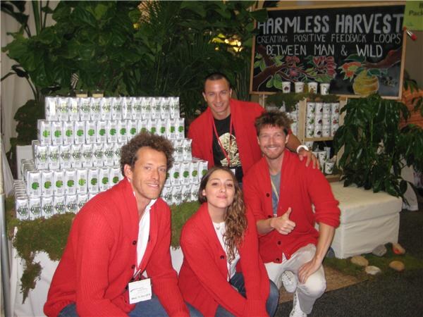Four people in red cardigans pose at a Harmless Harvest booth with stacked white cartons, moss, plants and a chalkboard sign; one man gives a thumbs up.