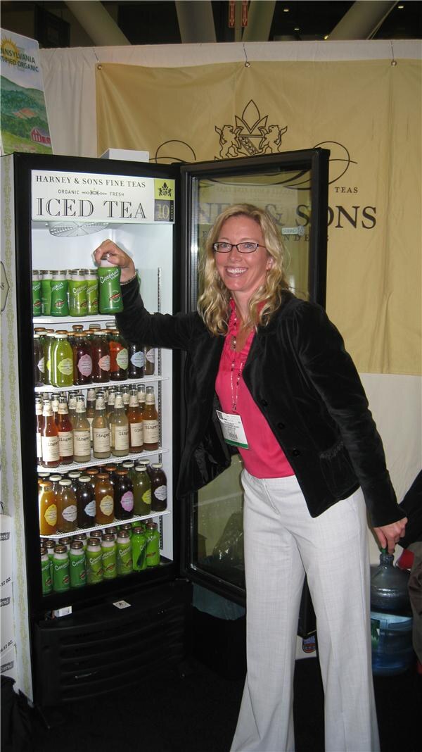 A smiling woman with blonde hair and glasses stands beside an open Harney & Sons Fine Teas Iced Tea refrigerator stocked with bottled teas, reaching for a green bottle on the top shelf.