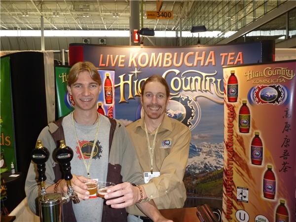 Two smiling men wearing badge lanyards hold small glasses of kombucha beside two tap handles, in front of a High Country Kombucha banner reading Live Kombucha Tea.