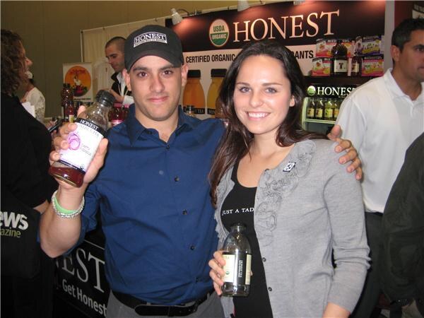 A man in a Honest cap holds up a bottle of Honest tea and a smiling woman holds another bottle, in front of a Honest USDA Organic sign at a trade-show booth.