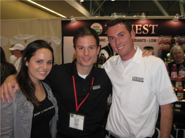 A woman and two men stand close together smiling in front of an Honest booth display, one man in a black shirt with a red lanyard and the other in a white Honest polo.