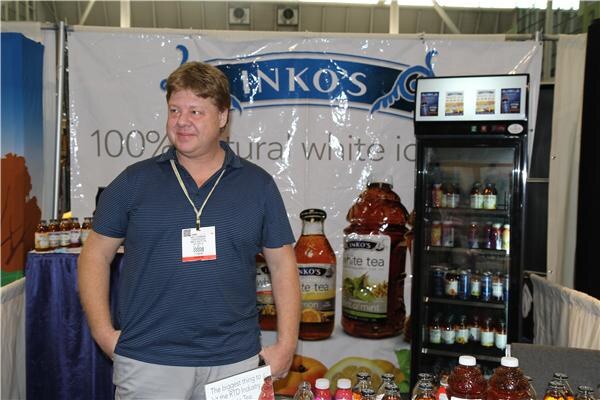 A man in a blue striped polo with a badge lanyard stands in front of an Inko's white tea banner, with bottled tea on a table and a lit refrigerated cooler beside him.