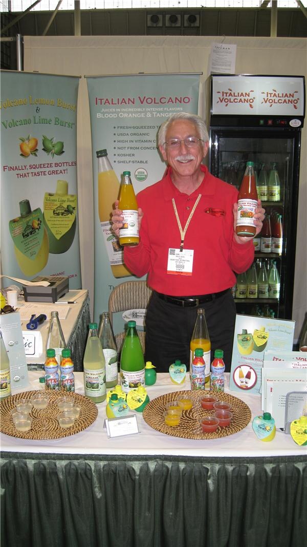 A gray-haired man in a red shirt holds two bottles of juice behind a table of bottles and samples, with Italian Volcano and Volcano Lemon Burst and Lime Burst banners and a refrigerator behind him.