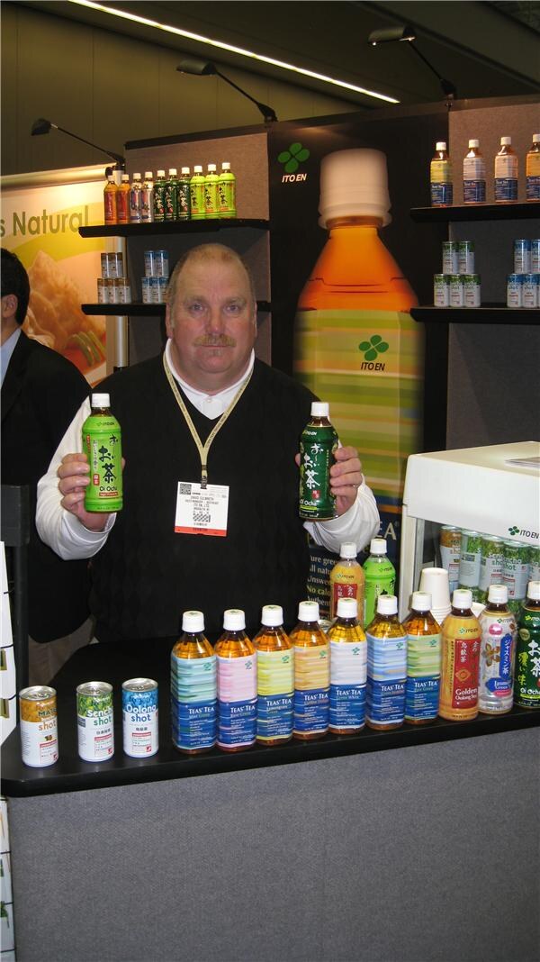 A mustached man with a badge lanyard holds up two green bottles of Ito En tea, with rows of bottled teas and canned shots on the counter and shelves behind him.