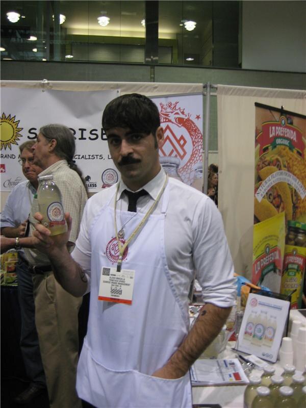 A man with a mustache, white apron and tie holds up a bottle of drink beside a La Preferida Organic banner, with other people standing behind him.