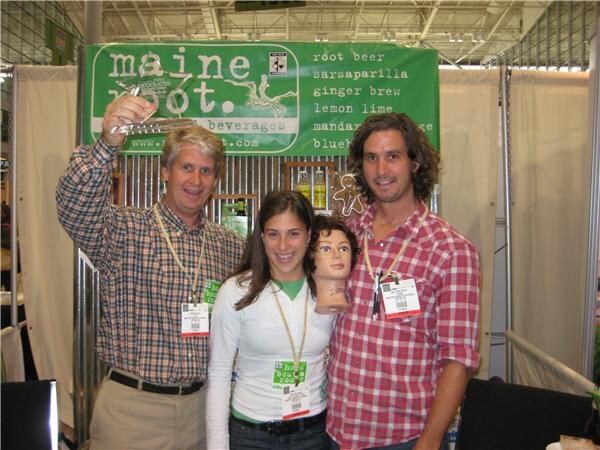 Two men and a woman wearing badges stand at a Maine Root booth; one man raises a glass trophy and the woman holds a mannequin head in front of the green Maine Root banner.