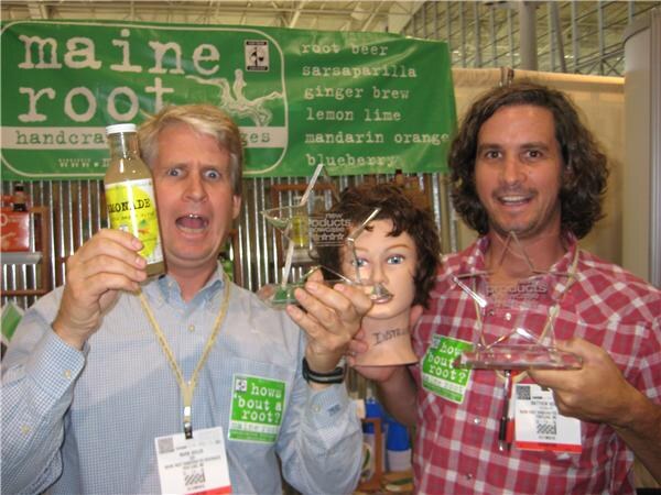 Two men with badges pose at a Maine Root booth, one holding a bottle of lemonade and glass awards, the other holding a glass award, with a mannequin head between them.