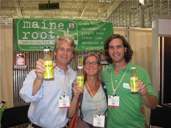 Two men and a woman with glasses hold bottles of Maine Root lemonade and smile in front of the green Maine Root banner listing root beer, sarsaparilla and other flavors.