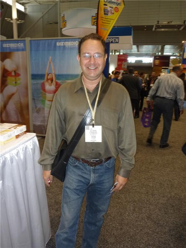 A smiling man with glasses, a lanyard badge and a shoulder bag stands in front of Enzymedica banners in a busy exhibit hall aisle.