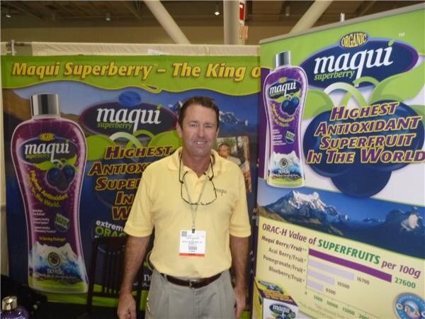 A man in a yellow polo with a badge stands in front of Maqui Superberry banners showing purple bottles and text about antioxidants and ORAC values.