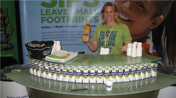 A woman in a green T-shirt stands behind a glass counter holding a Naked juice bottle, with rows of Naked cartons in front of her and a recycling sign behind.