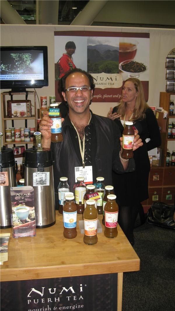 A smiling man with glasses holds two bottled Numi iced teas at a wooden counter with more bottles and a Numi Puerh Tea sign; a woman stands behind him.