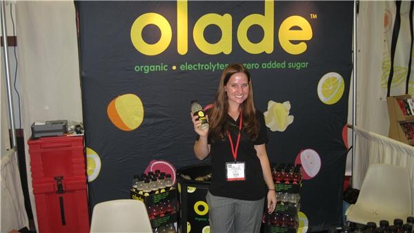 A woman with long brown hair holds a bottle in front of a black Olade banner, with cases of Olade bottles beside her.