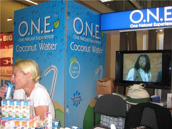 A blonde woman drinks from a carton next to a blue O.N.E. Coconut Water display, with boxes of cartons in front and a television on the right.