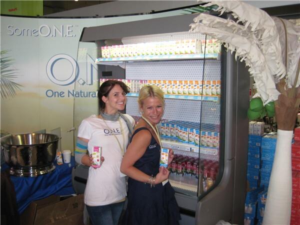 Two smiling women in O.N.E. shirts each hold a small carton beside a refrigerated display case stocked with cartons, with an O.N.E. sign and a decorative palm tree nearby.
