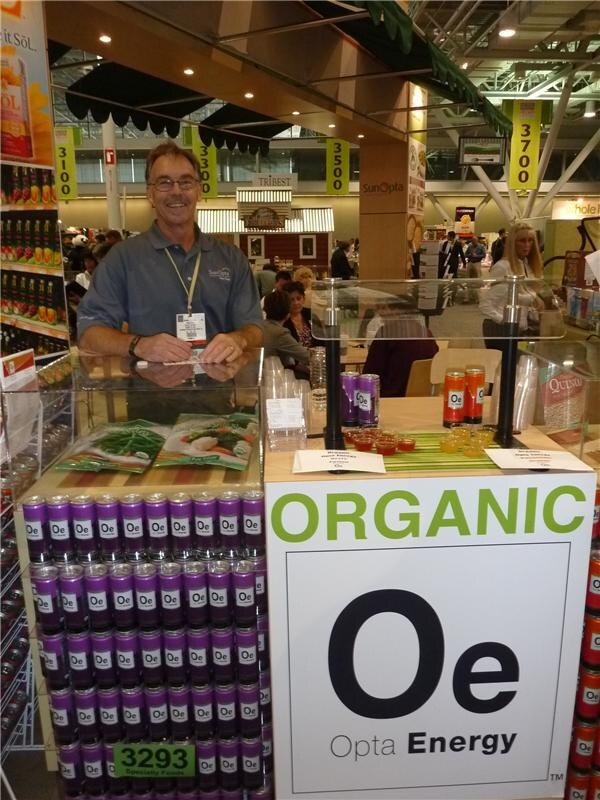 A smiling man with glasses and a badge leans on a glass counter at the SunOpta booth. Purple Opta Energy cans are stacked below, and a sign reads "Organic Oe Opta Energy."