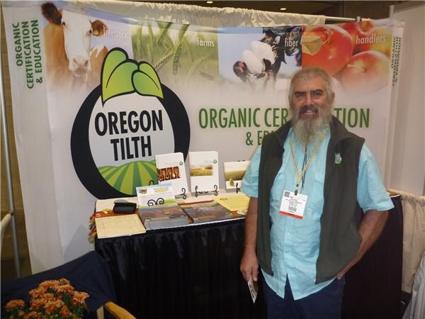 A bearded man in a light blue shirt and dark vest stands beside a table of brochures in front of an Oregon Tilth organic certification banner.