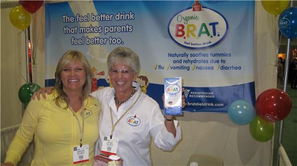 Two smiling women with badges stand in front of an Organic B.R.A.T. banner; one holds up a carton of the drink. Colorful balloons surround the booth.