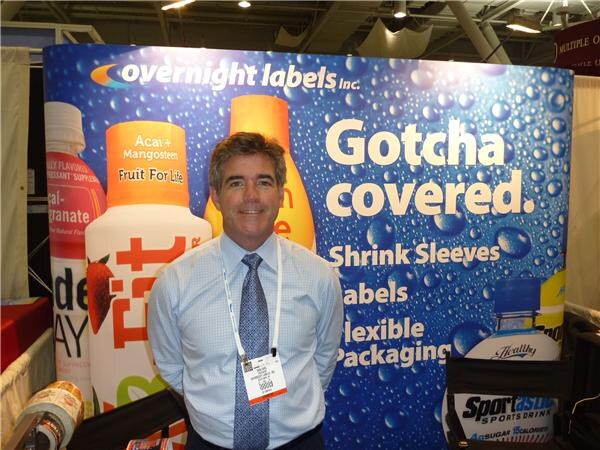 A man in a blue dress shirt and tie stands in front of an Overnight Labels Inc. backdrop reading "Gotcha covered" with shrink sleeves, labels and flexible packaging.