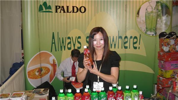 A woman in black holds a bottle of aloe drink in front of a green Paldo banner, with rows of aloe bottles on the table and a man in a white shirt behind her.