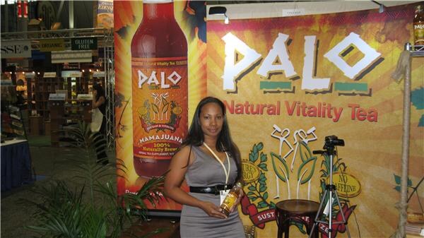A woman in a gray dress and lanyard holds a bottle of Palo tea in front of a Palo banner reading Natural Vitality Tea, with a large Mamajuana bottle graphic beside her.